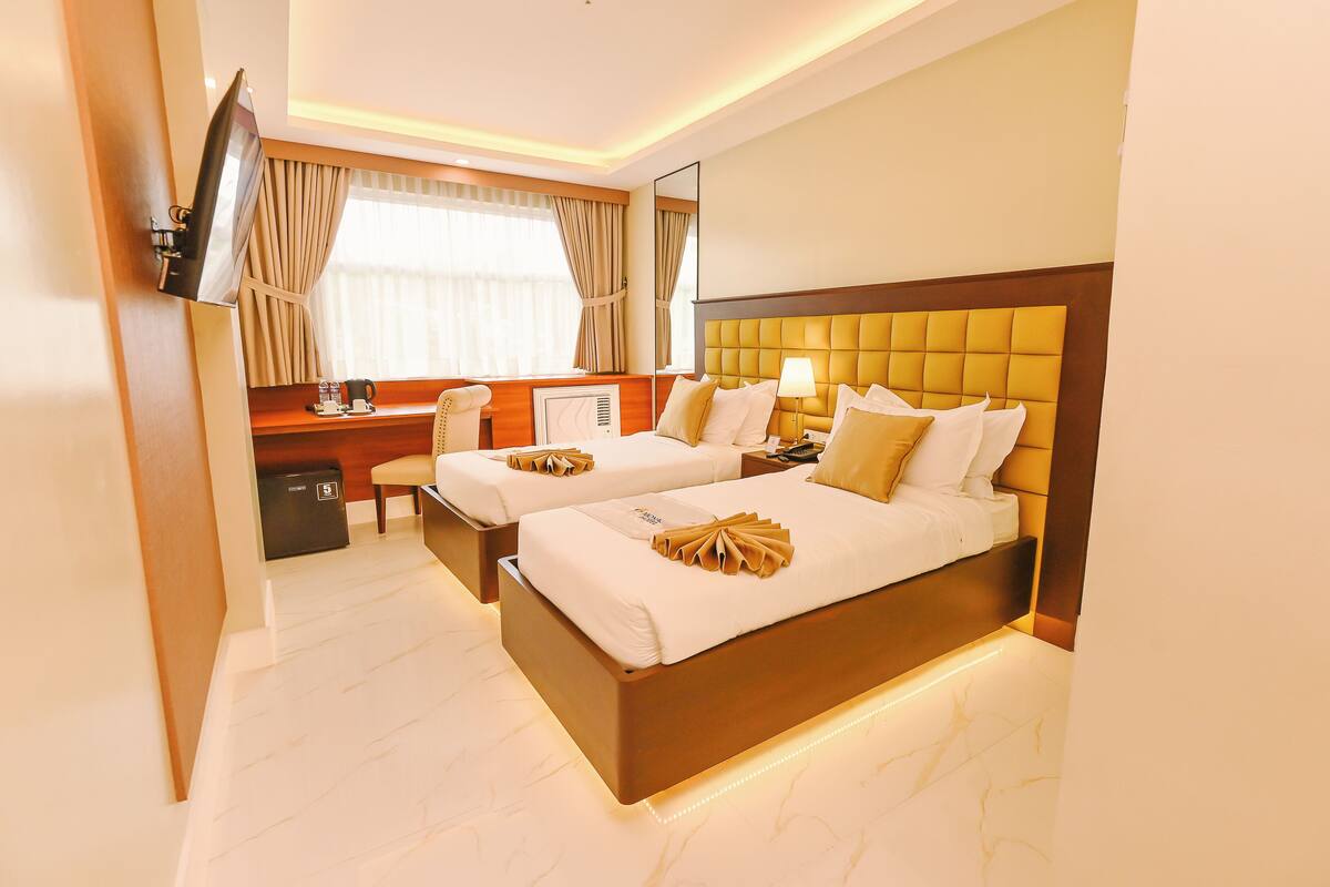 A hotel room with two beds, a desk, a chair, a TV, and a window with curtains.