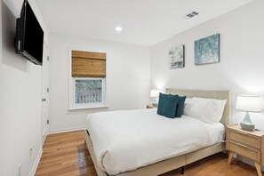 A bedroom with a bed, bedside tables, a TV, a window with blinds, and artwork on the wall.