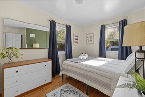 Ett sovrum med en säng, byrå, spegel, fönster med gardiner, en lampa och en liten växt.