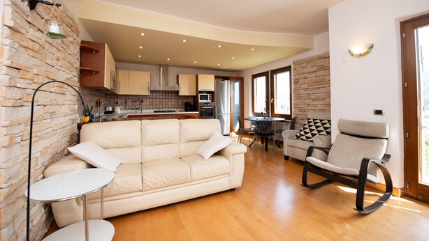 Een moderne woonkamer met een beige bank, een schommelstoel en een kleine tafel. In de achtergrond is een keuken zichtbaar met houten kasten en een stenen muur.
