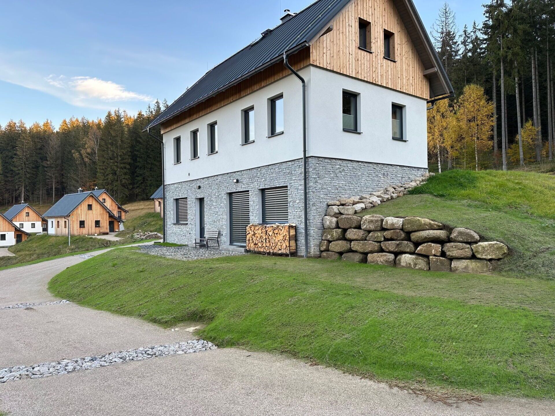 A modern two-story building with a stone facade and wooden upper levels, surrounded by a rural landscape with a gravel path leading to the entrance.