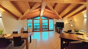 Ein geräumiges Loft mit hohen Decken und freiliegenden Holzbalken. Es bietet einen Essbereich mit Tisch und Stühlen, eine Sitzecke mit Sofa und Fernseher sowie einen Balkon mit Blick.