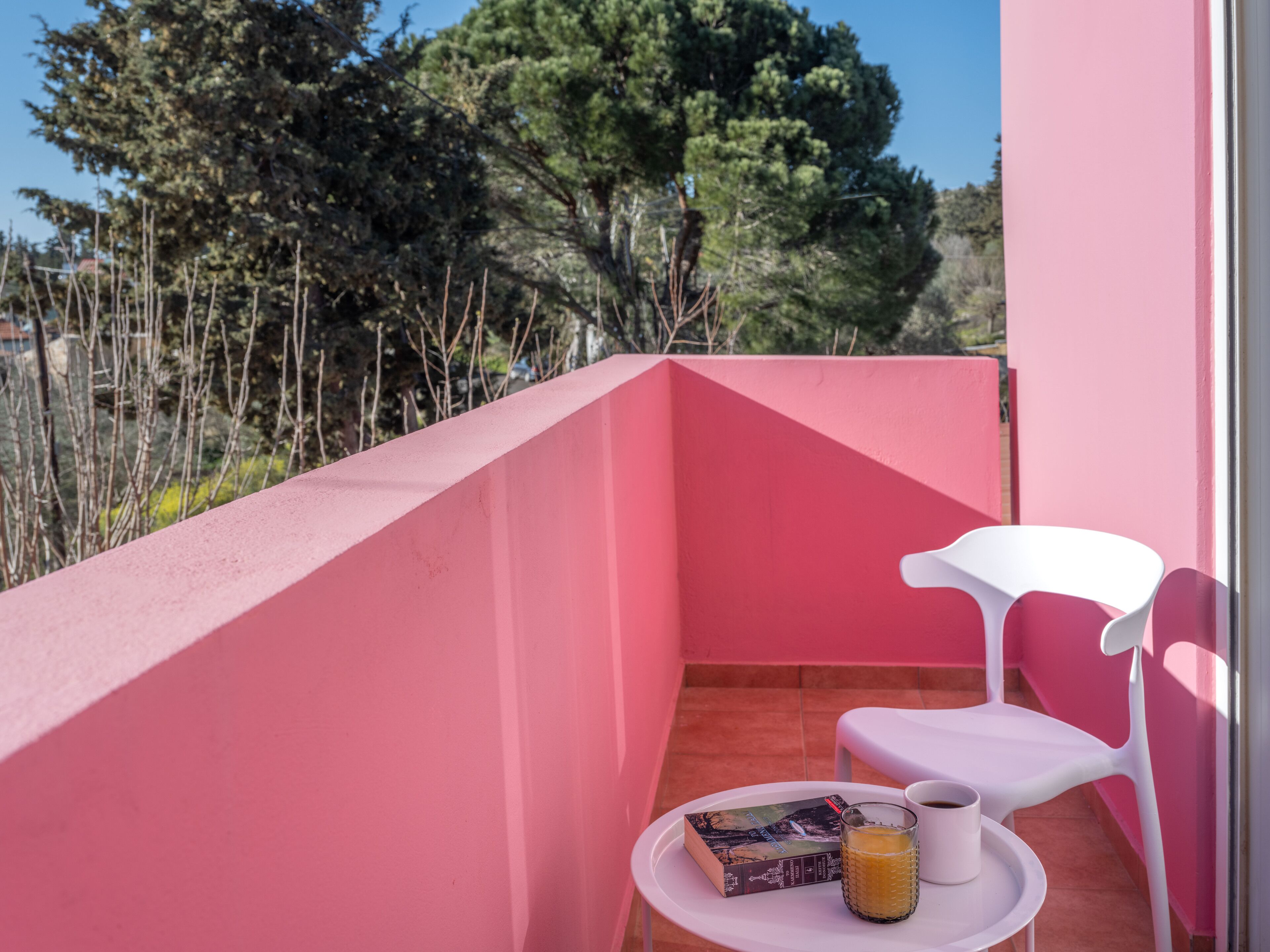A balcony with a pink wall, a white chair, a small table, a book, and a glass of orange juice.
