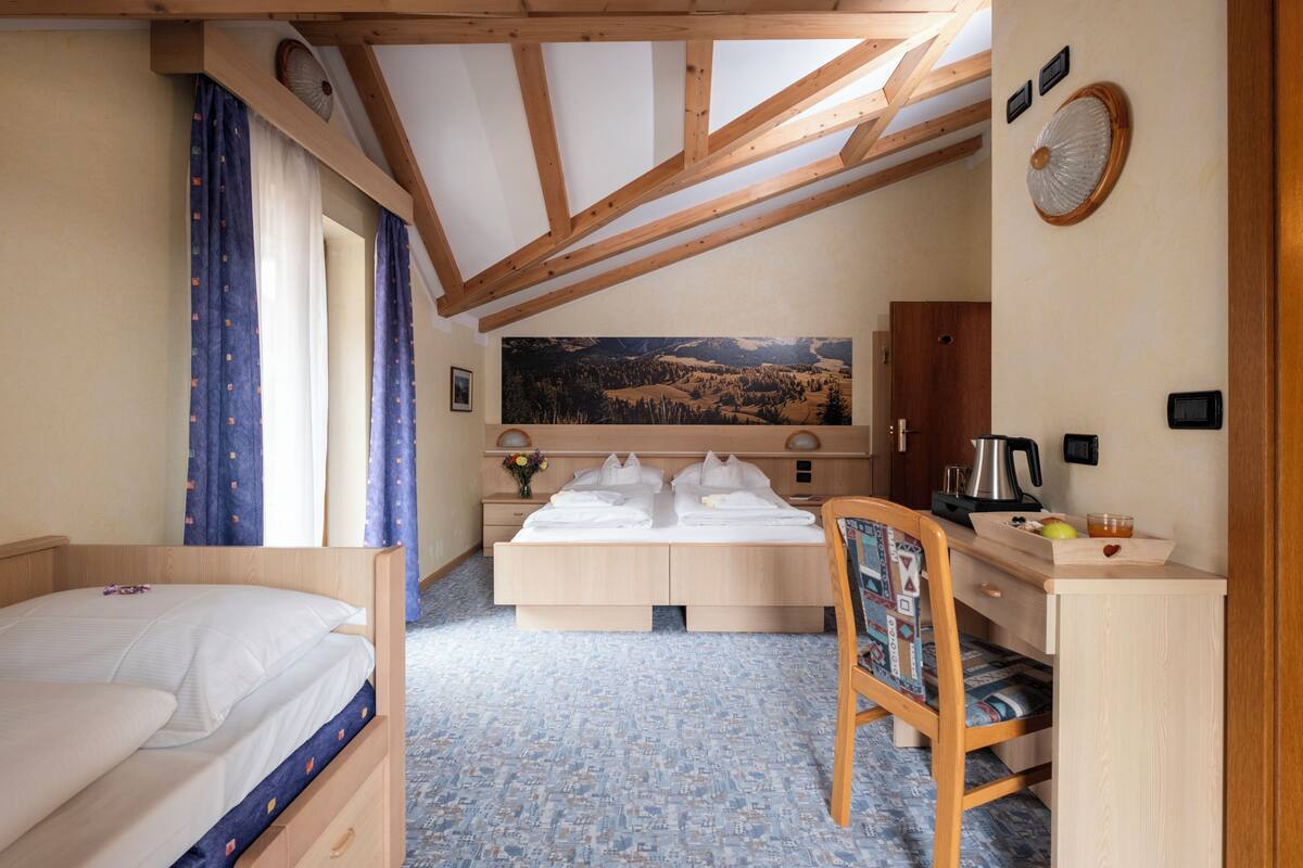 Een kamer met een bed, een bureau met stoel, een groot schilderij en een raam met gordijnen.
