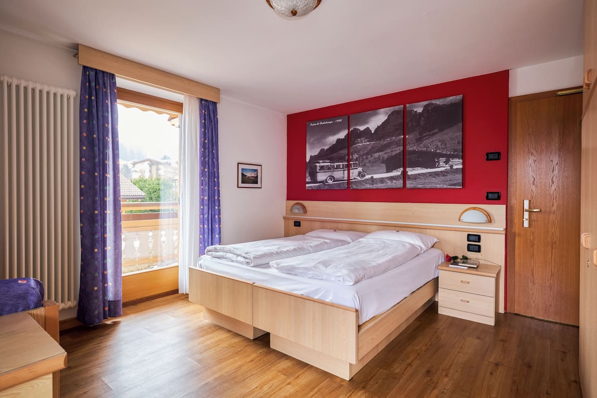 Een slaapkamer met een bed, houten vloer, een raam met gordijnen, een rode muur met een schilderij en een deur.