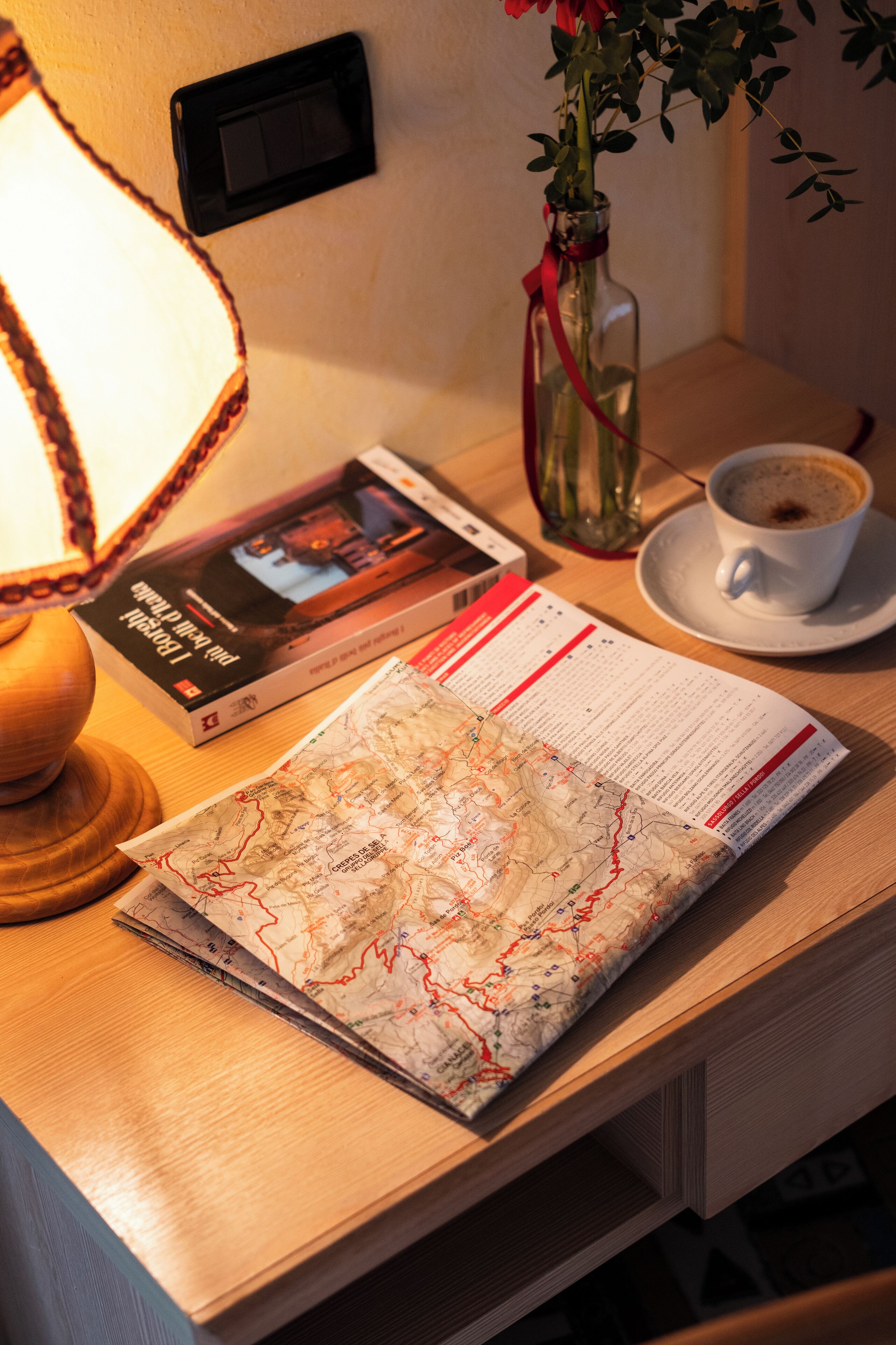 Une table en bois sur laquelle se trouvent une carte, un livre, un vase avec des fleurs, une tasse de café et une lampe.