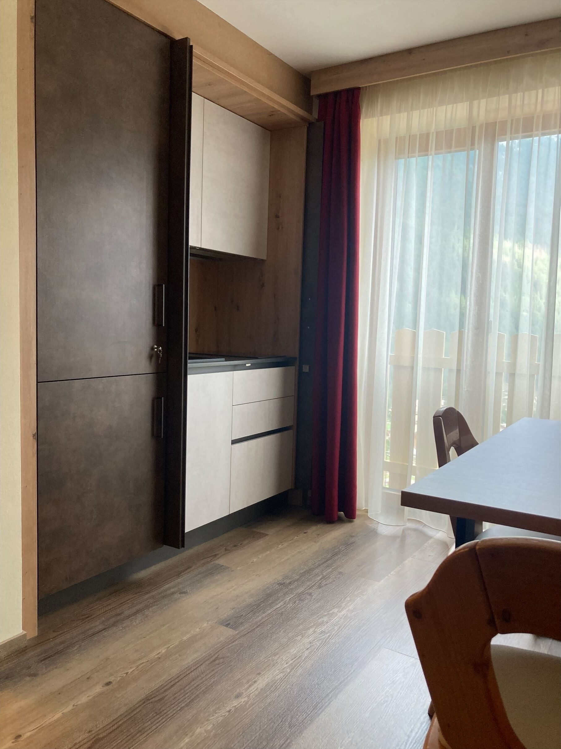 Ein Zimmer mit Holzboden, einem Tisch, einem Stuhl, einem Schrank und einem Vorhangfenster.