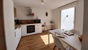 Et moderne køkken med hvide skabe, en træbordplade og en indbygget ovn. Der er et spiseområde med et hvidt bord og stole, og et stort vindue giver naturligt lys.