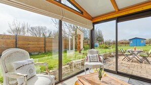 Une véranda dotée d’une porte vitrée donnant sur un patio. À l’intérieur, on trouve deux fauteuils blancs recouverts de tissu, agrémentés d’un coussin, une table basse en bois sur laquelle est posé un vase de fleurs, et une vue sur un abri de jardin bleu situé dans le jardin.
