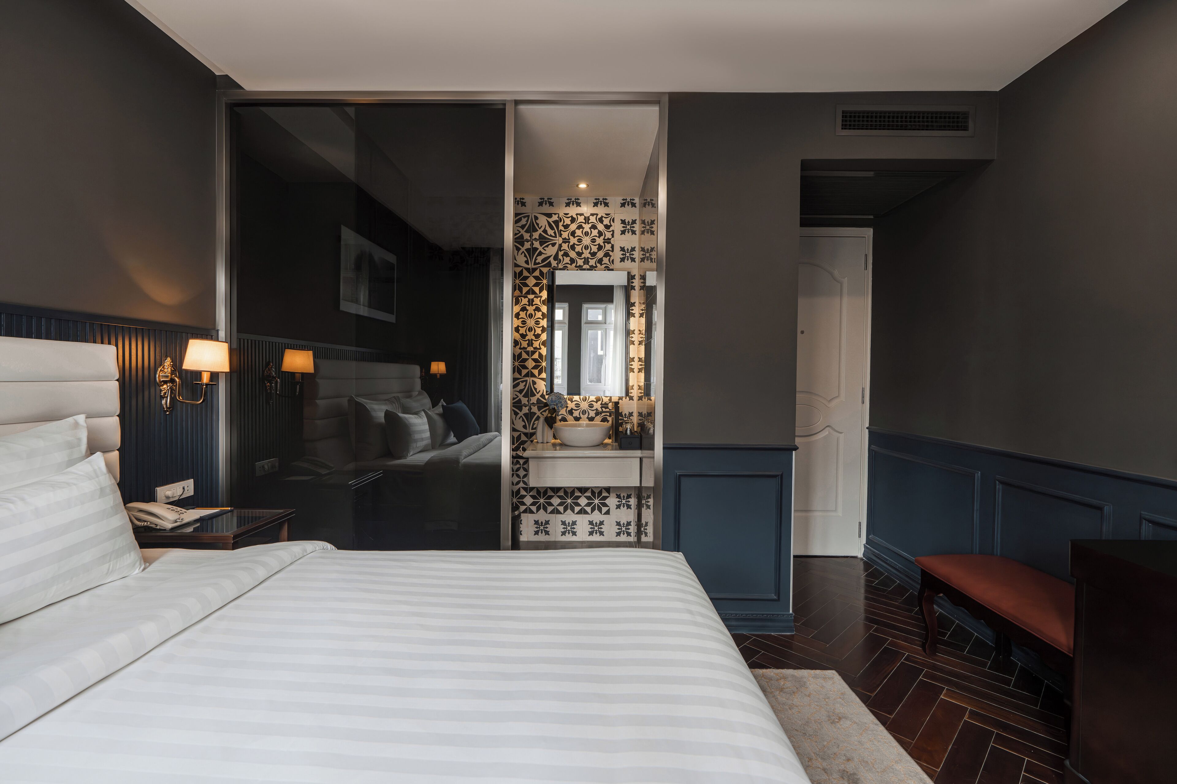 A modern hotel room with a large bed, a small desk, a bench, and a bathroom visible through an open door.