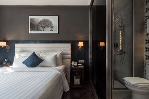 Une chambre d’hôtel avec un grand lit, une table de chevet avec une lampe, une petite table avec un téléphone et une lampe, et un espace douche.