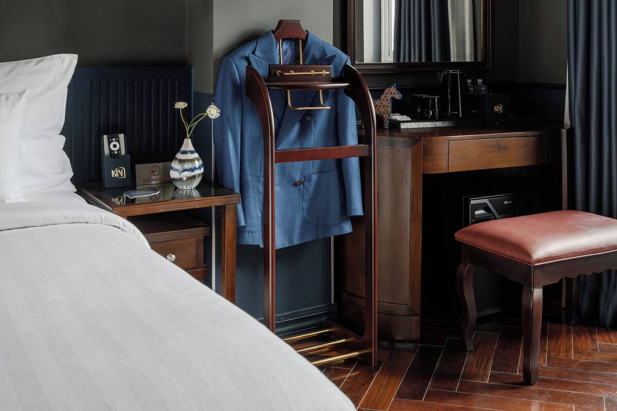 Una habitación de hotel con una cama, un escritorio con espejo, una silla, una mesita con un jarrón y una lámpara colgante.