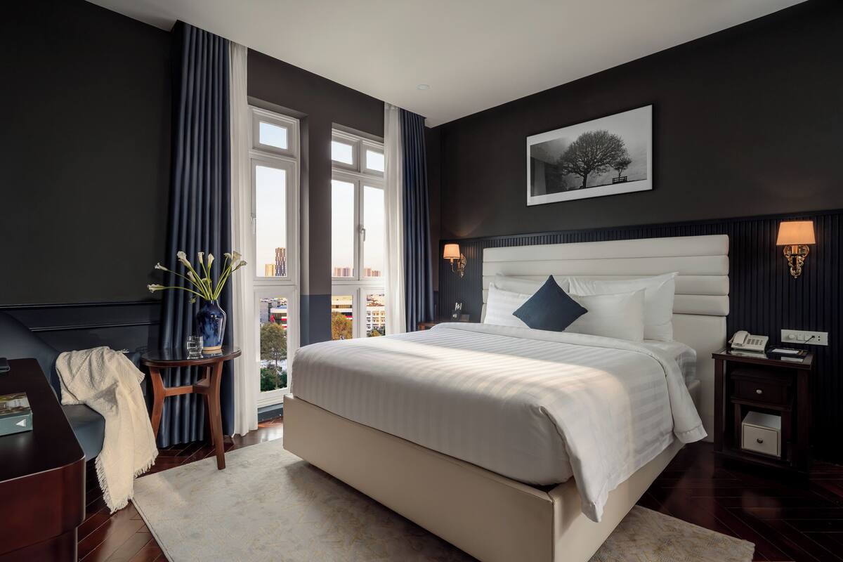 Un dormitorio moderno con una cama grande, una mesita de noche con lámpara, una pequeña mesa con un florero y una ventana con cortinas.