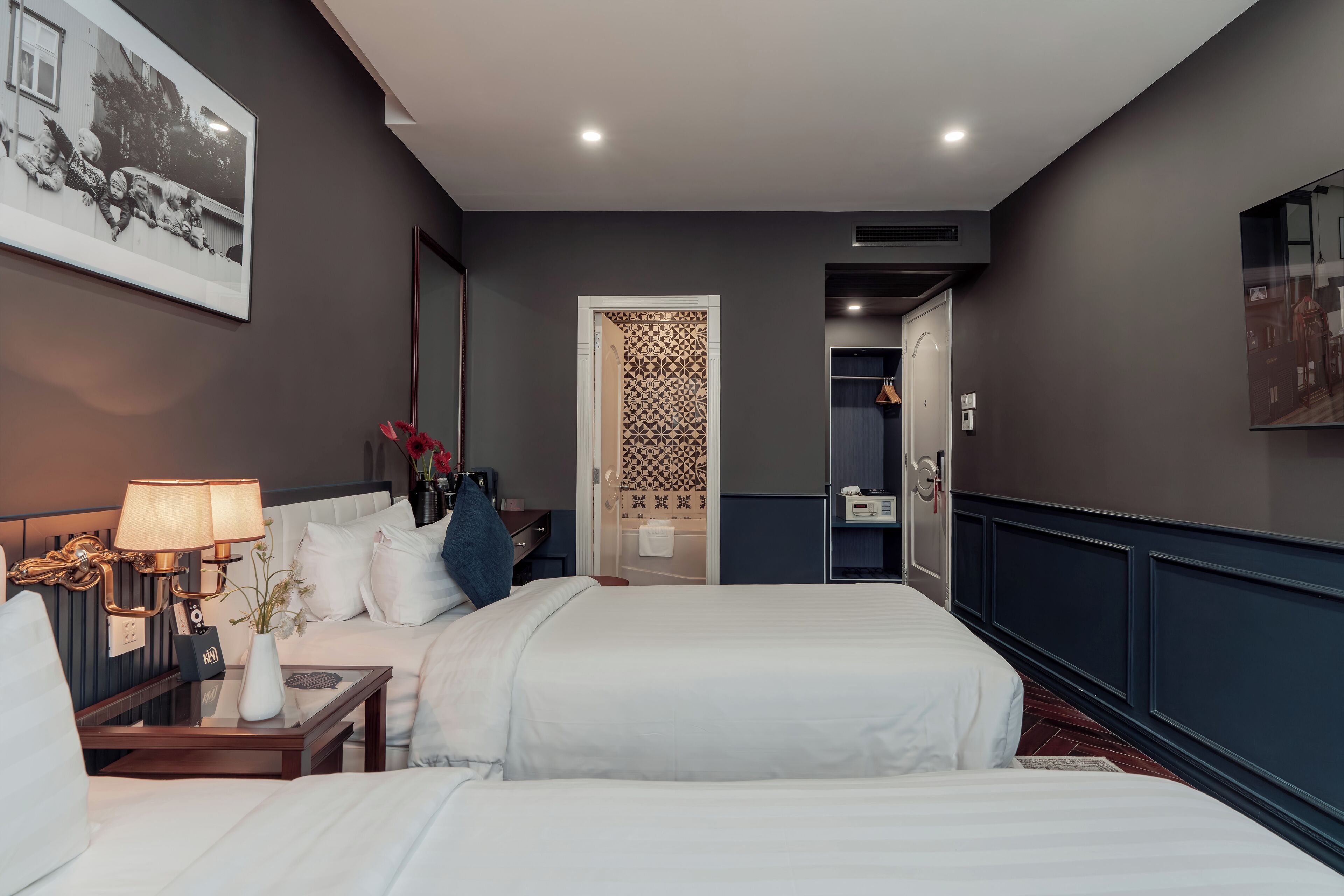 Ein Hotelzimmer mit zwei Betten, einem Schreibtisch mit Lampe, einer Vase mit Blumen und einem gerahmten Bild an der Wand.