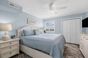 Quarto com cama, mesas de cabeceira, cômoda, ventilador de teto, janela e porta.