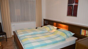 A double bed with a checkered bedspread, a bedside table with books, and a window with curtains. - Ferienwohnung Miezal, 1-4 Pers (Raesfeld)