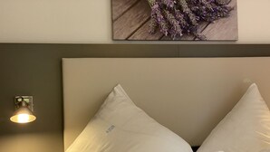 A neatly made bed with white linens, a wall-mounted light fixture, and a framed picture of flowers on the wall.