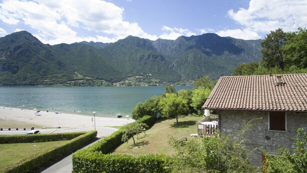 A stone building with a tiled roof, a beach, and mountains in the background.