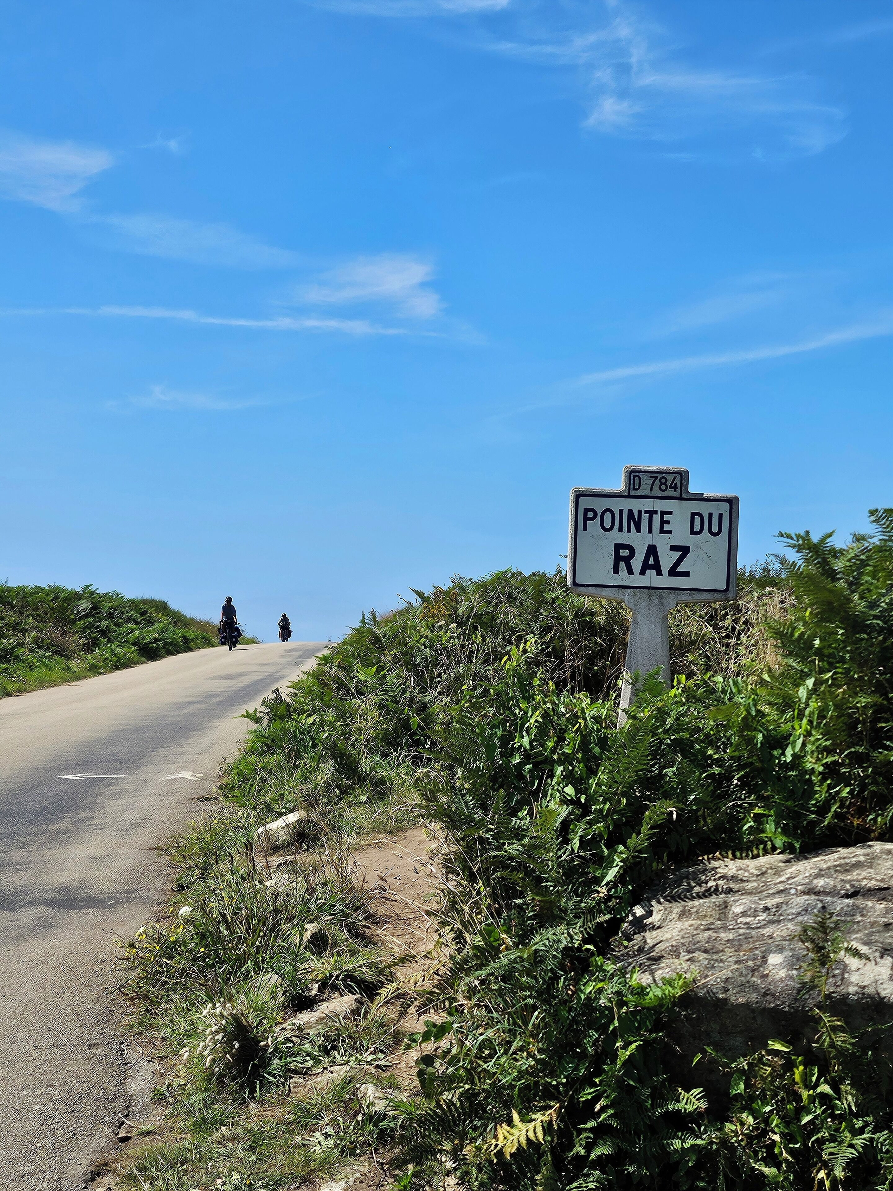 Tienvarressa on kyltti, jossa lukee ”POINTE DU RAZ”, taivaalla on kirkas sininen taivas ja kauempana näkyy moottoripyöräilijä.