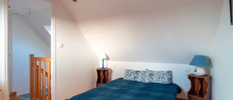 A bedroom with a wooden ceiling, a bed with a blue bedspread, two bedside tables with lamps, and a wooden staircase.