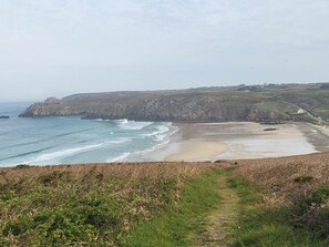 Un paisaje costero con una playa arenosa, acantilados rocosos y un sendero cubierto de hierba que conduce a la orilla.