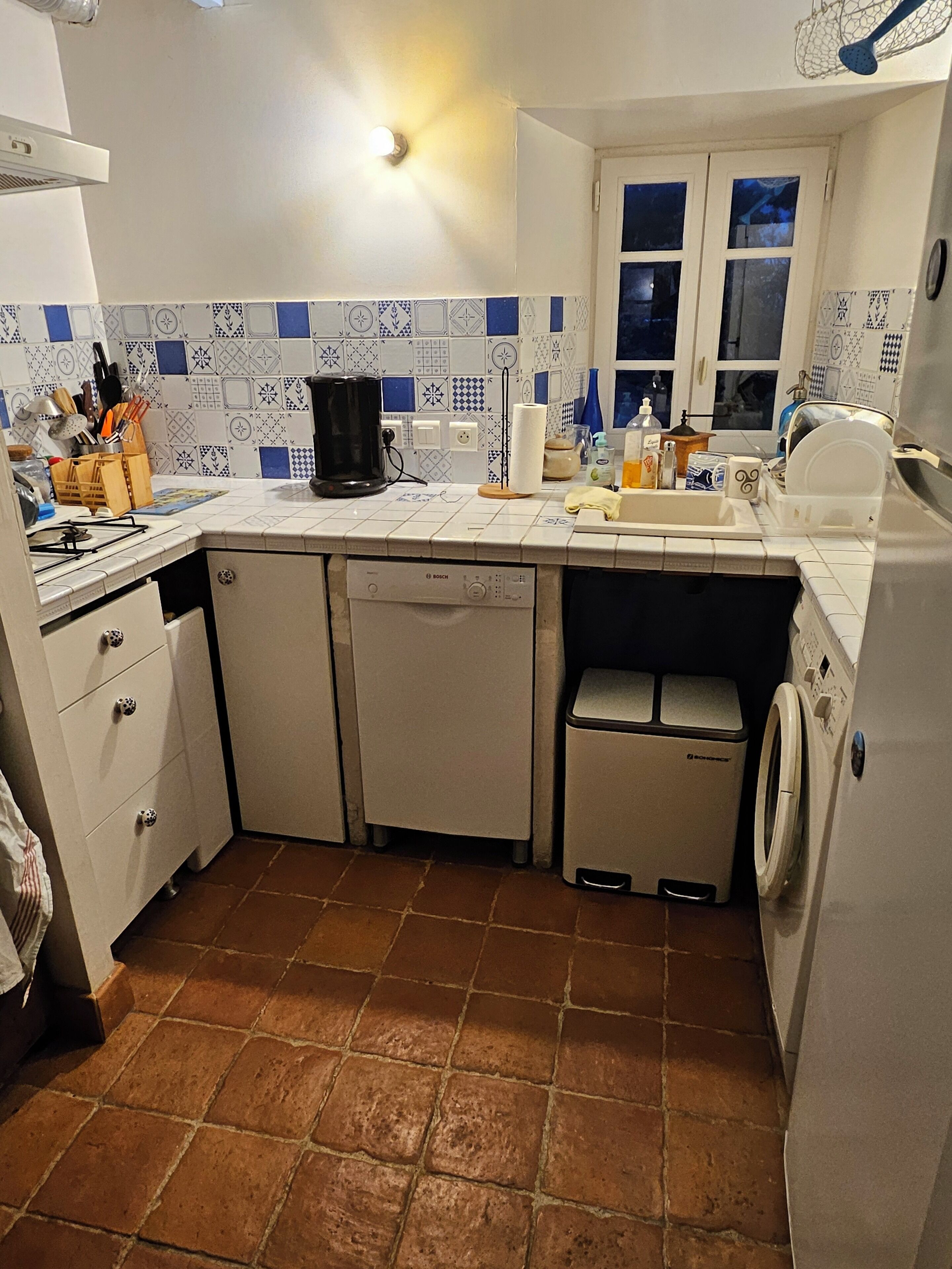 Une cuisine compacte avec des meubles blancs, un lave-vaisselle Bosch, un lave-linge et une fenêtre offrant une belle vue.