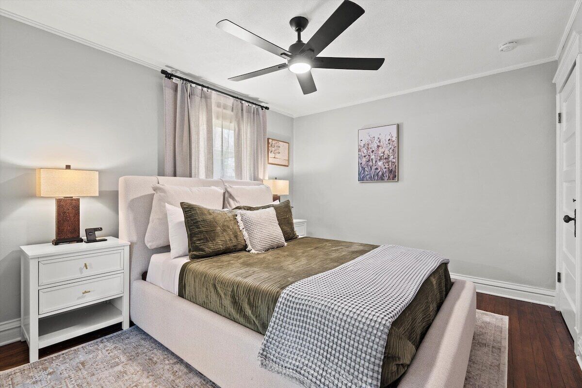 A bedroom with a bed, nightstand, lamp, ceiling fan, and artwork on the wall.