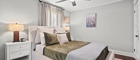 Un dormitorio con cama, mesita de noche, lámpara, ventilador de techo y una obra de arte en la pared.