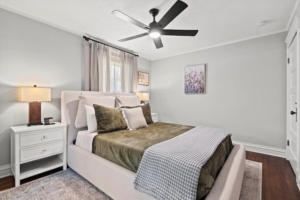 Un dormitorio con cama, mesita de noche, lámpara, ventilador de techo y una obra de arte en la pared.