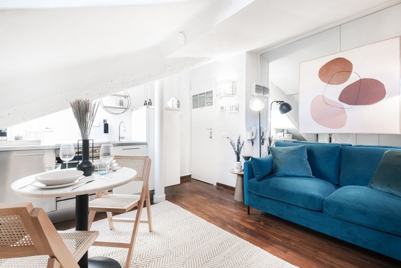 A modern living room with a blue sofa, a dining table with chairs, and a kitchen area in the background.