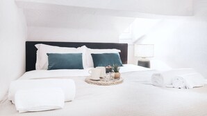 Un lit bien fait, agrémenté d’oreillers blancs et turquoise, une table de chevet avec une lampe, une petite plante et un plateau avec une tasse, des lunettes et une bouteille.