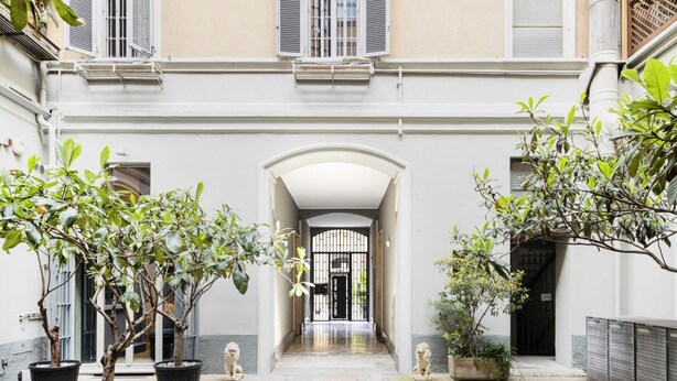 Une cour intérieure agrémentée d’arbres en pot, une arche centrale et la façade d’un bâtiment avec des volets.