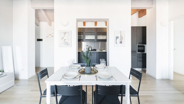 Un comedor moderno con una mesa blanca dispuesta para cuatro personas, rodeada de sillas negras. Se observa una cocina en el fondo con armarios oscuros y electrodomésticos de acero inoxidable.