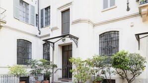 Un edificio con fachada blanca, ventanas amplias con rejas metálicas y una entrada central con escalones. Hay plantas en macetas a lo largo de la fachada y una barandilla metálica a la izquierda.