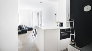 Una cocina moderna con armarios blancos, un horno negro y una encimera blanca. Se observa un reloj de pared negro y una estantería blanca con un florero y una cafetera.