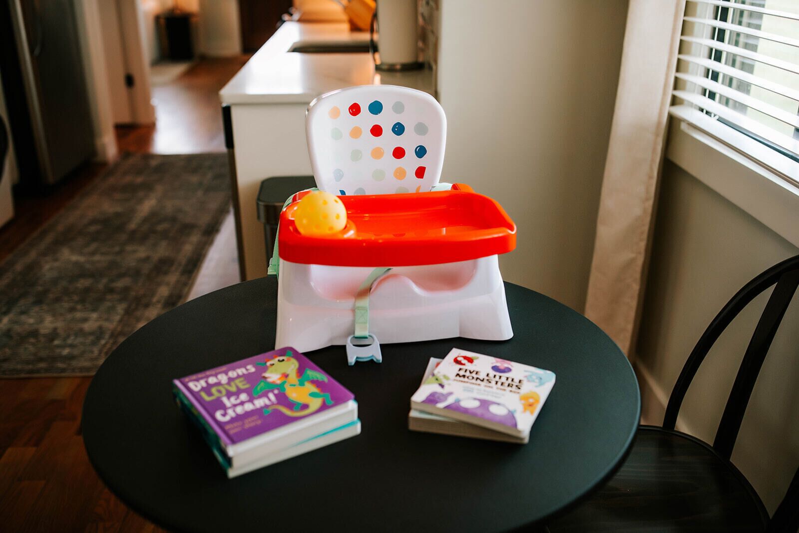 Une chaise haute pour bébé avec un plateau, deux livres et une fenêtre avec des stores.