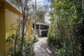 Uma trilha de pedra estreita conduz a uma área externa coberta, com uma estrutura branca, rodeada por vegetação densa e um edifício amarelo.