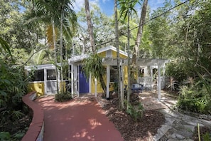 Um jardim com um caminho que leva a um edifício amarelo, com porta azul, cercado por árvores e plantas.