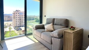 En moderne stue med en grå sofa, et lille træbord og et stort vindue med udsigt til by og grønne områder.