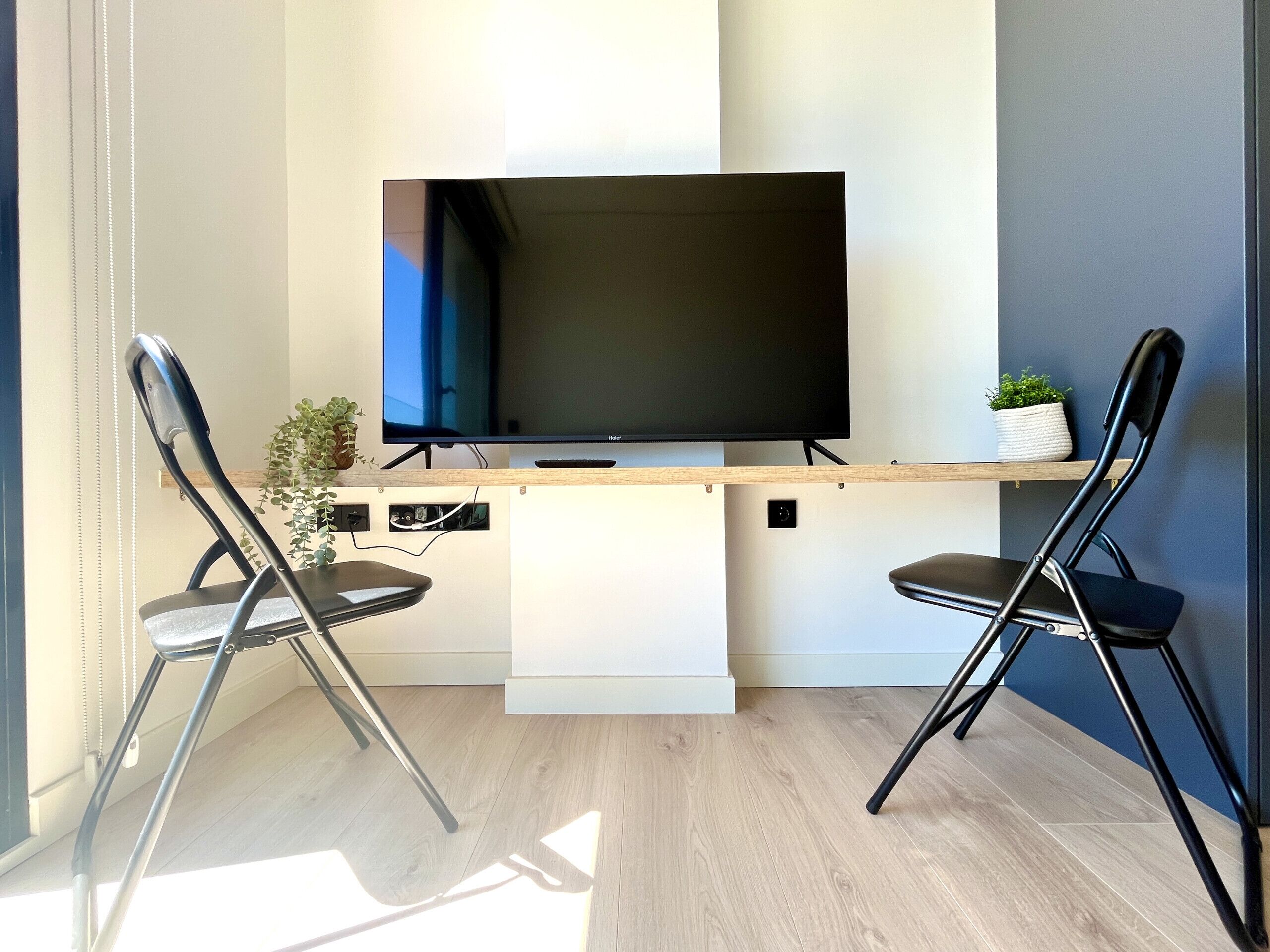 Ein modernes Wohnzimmer mit einem Flachbildfernseher auf einem Holzständer, zwei schwarzen Sesseln, einer kleinen Pflanze und einem weißen Regal.