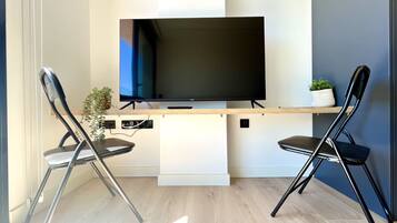 Ein modernes Wohnzimmer mit einem Flachbildfernseher auf einem Holzständer, zwei schwarzen Sesseln, einer kleinen Pflanze und einem weißen Regal.