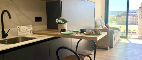 Una cocina moderna con pileta integrada, refrigerador, comedor con mesa y sillas, y vista de la ciudad a través de grandes ventanales.