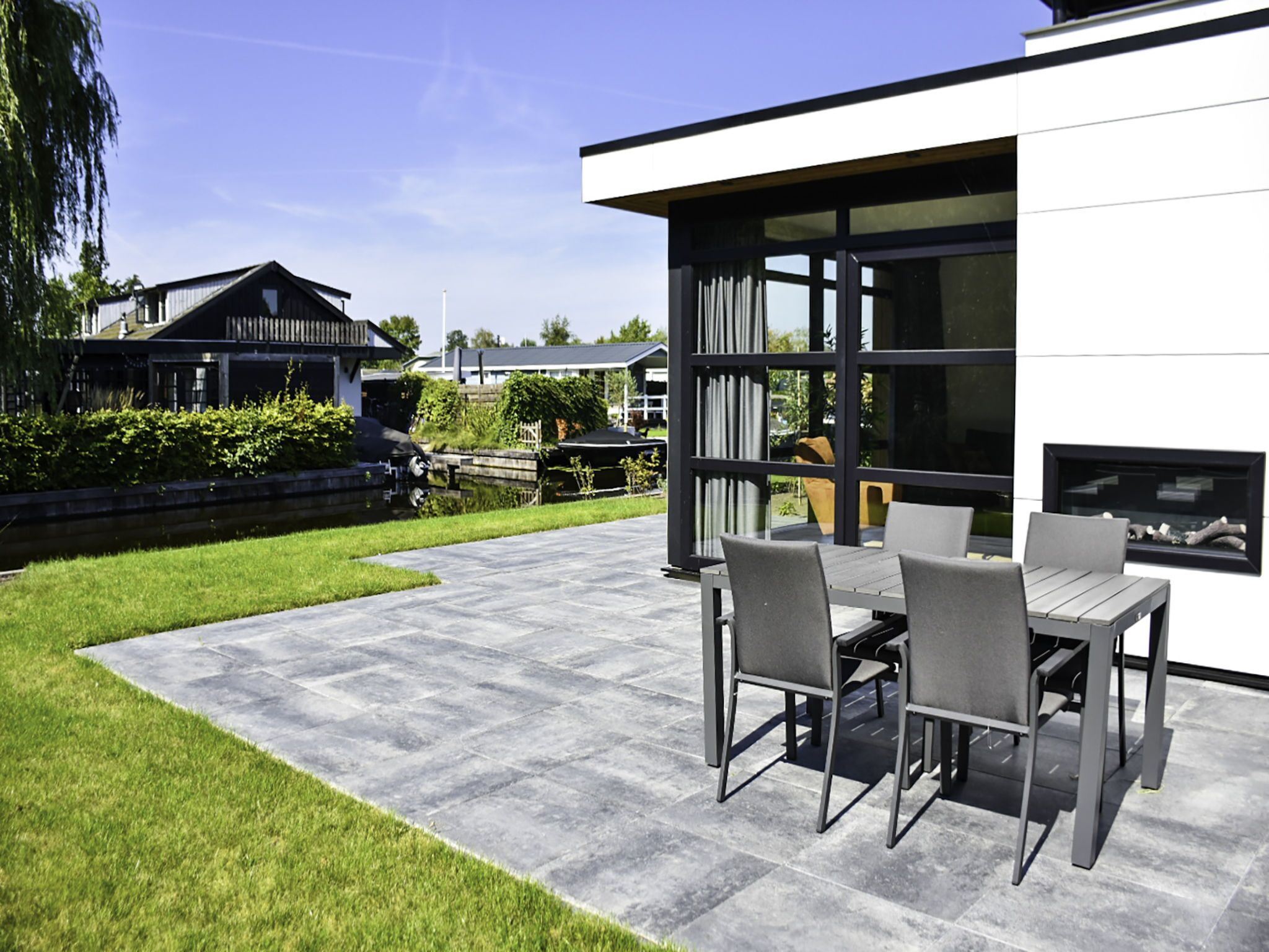 A modern outdoor patio with a stone floor, a dining table with chairs, and a large window overlooking a garden.