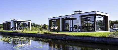A modern houseboat with a glass facade, a small deck, and a water body in the foreground.