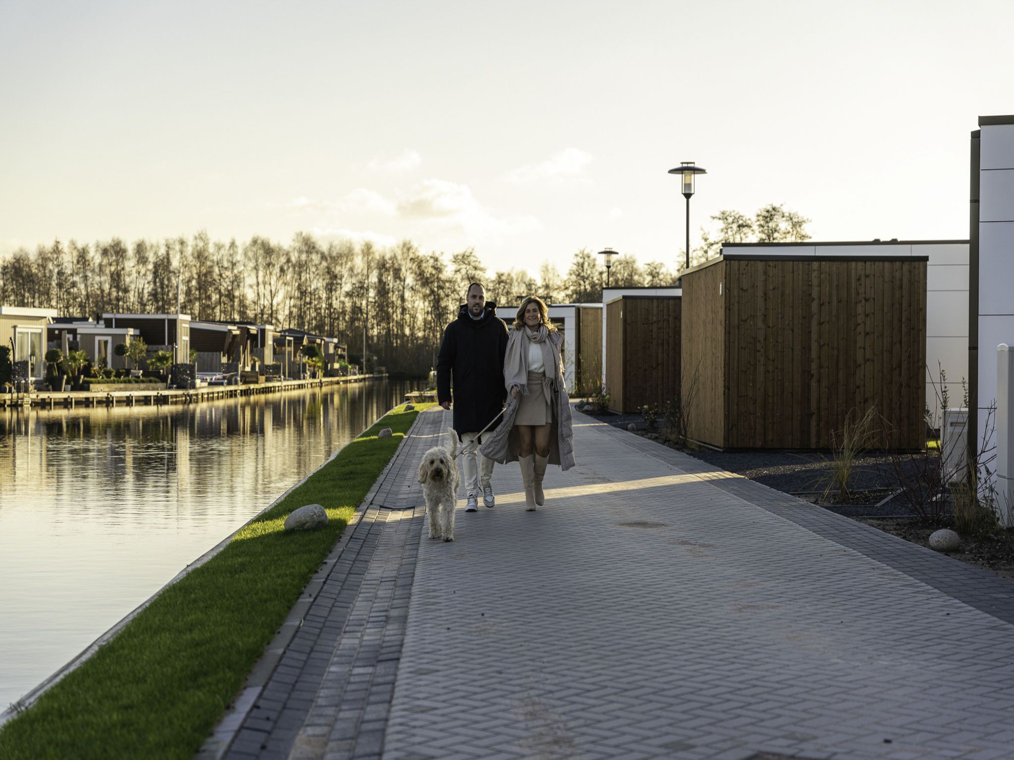 Een pad langs een waterpartij, met moderne gebouwen en een houten hek op de achtergrond.