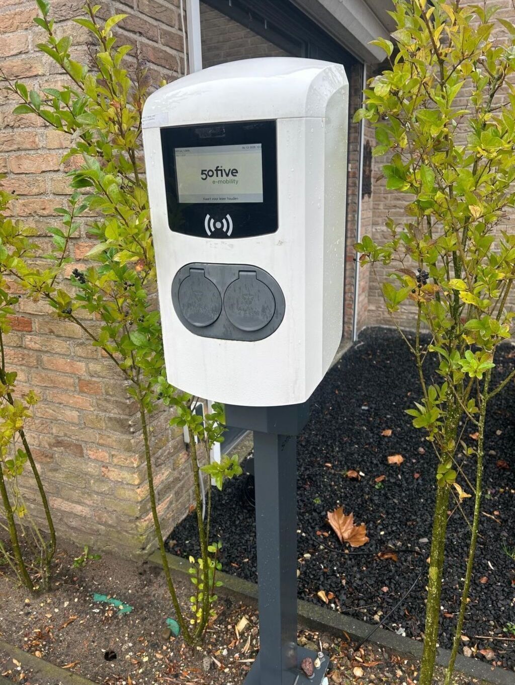 Una estación de carga pública con la marca '50five e-mobility', con dos puntos de carga, situada al aire libre cerca de un edificio de ladrillo y rodeada de vegetación.