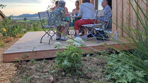 En grupp människor sitter vid ett utomhusbord på en träveranda, omgivna av grönska och med utsikt över landskapet.