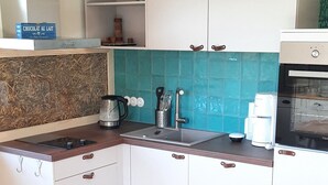 Ett kompakt kök med vita skåp, en träbänkskiva och ett grönt stänkskydd. På bänkskivan står en kaffebryggare, en vattenkokare och en brödrost. En vit stol är placerad vid ett litet runt bord.