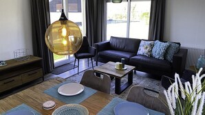 A modern living room with a sofa, dining table, and chairs. There is a large pendant light, a vase with flowers, and a view of the outside through large windows. - De Noordzee by Interhome (Scherpenisse)
