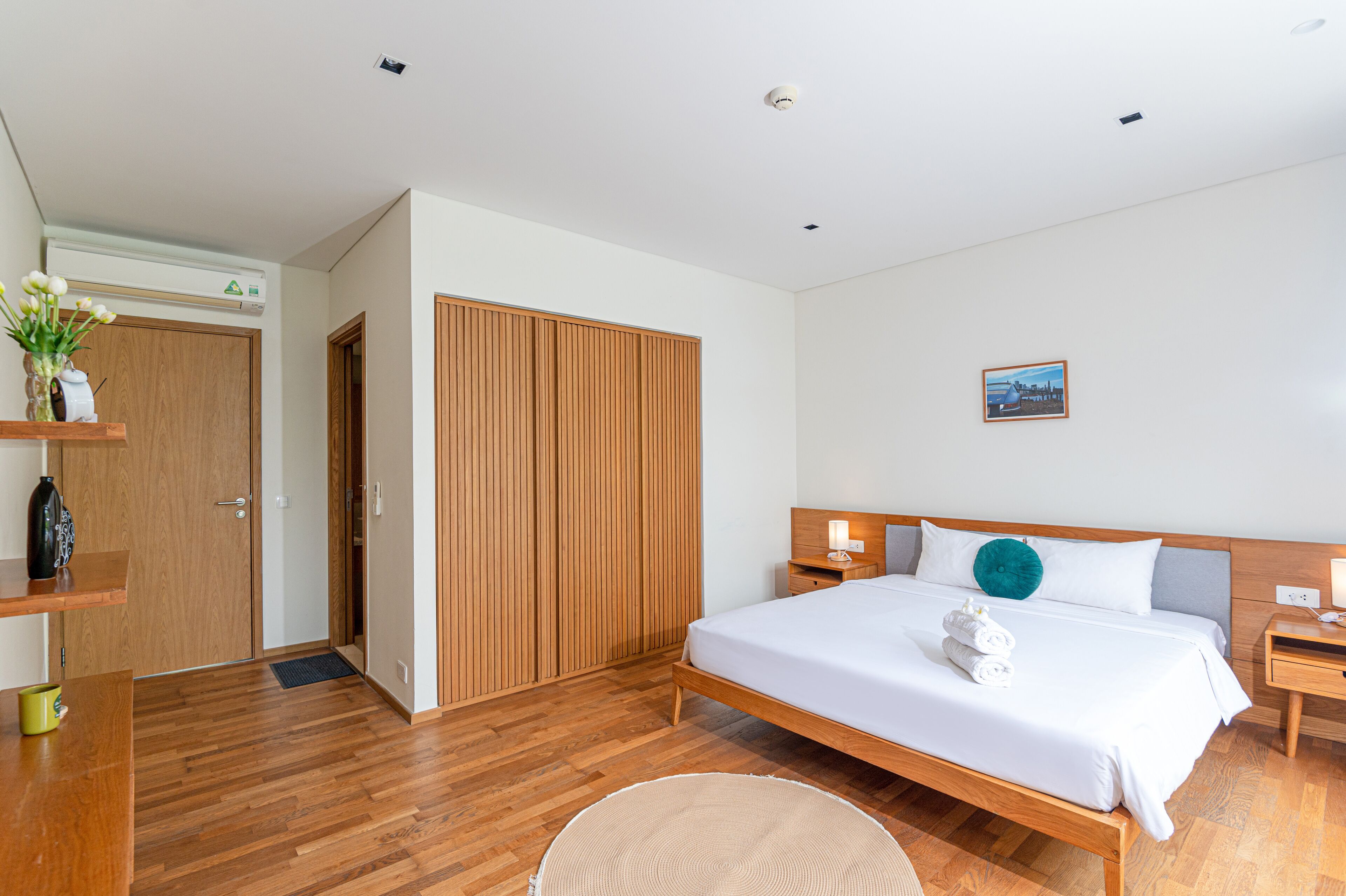 Une chambre d’hôtel moderne avec un lit en bois, des tables de chevet, une petite table de nuit, un tableau accroché au mur et une armoire en bois.